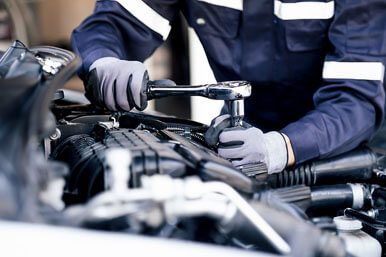 A close-up of an auto mechanic wearing a blue jumpsuit and gloves holding a wrench while doing a repair on a car engine.