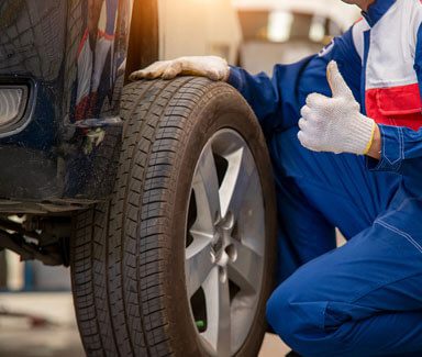 A close-up of an auto mechanic removing a car tire while doing a thumbs-up gesture.