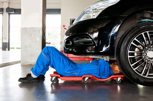 A wide shot of an auto mechanic in a blue jumpsuit lying on a creeper tool under a black car.