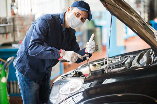 A wide shot of an auto mechanic in a dark blue jacket wearing gloves, a mask, a cup, and jeans while holding a wrench and doing some repairs on a black car engine.